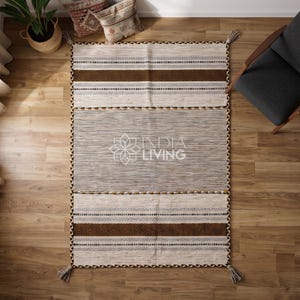 May include: A rectangular area rug featuring a central gray section flanked by cream-colored panels with brown and black geometric striped patterns. The rug has braided edges and tassels at the corners, with the text INDIA LIVING printed in the center.