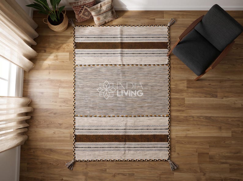 May include: A rectangular area rug featuring a central gray section flanked by cream-colored panels with brown and black geometric striped patterns. The rug has braided edges and tassels at the corners, with the text INDIA LIVING printed in the center.