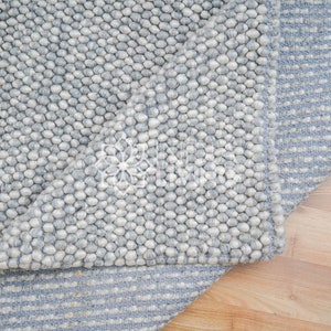 May include: A gray and white wool rug with a textured, bumpy surface. The rug has a striped pattern on the bottom edge.