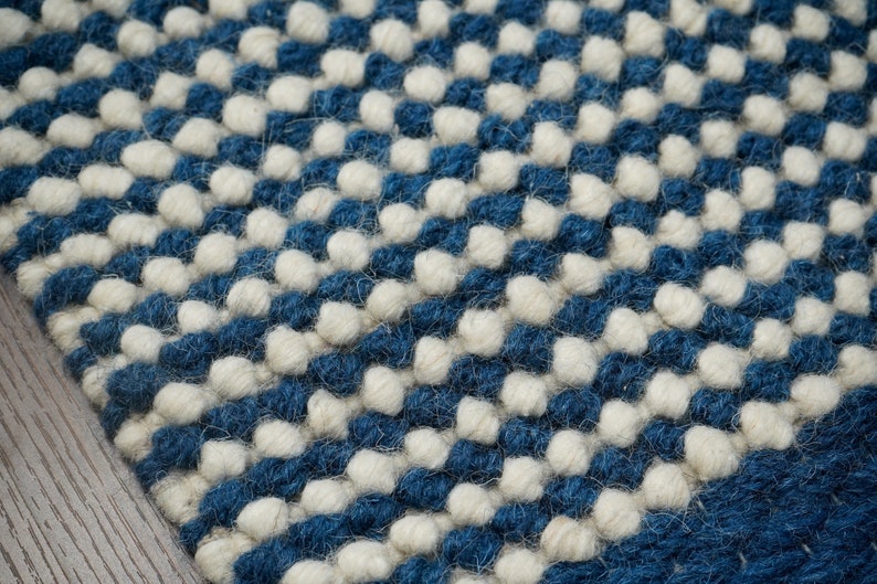 May include: Close-up of a blue and white woven rug with a textured, bumpy surface. The rug is made of wool and has a modern, geometric design.