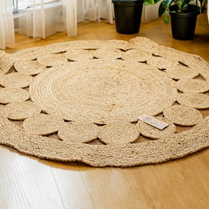 May include: A round, natural fiber rug with a woven design. The rug has a circular pattern of cutouts around the edge, creating a decorative border. The rug is made of natural jute fibers and has a light brown color.