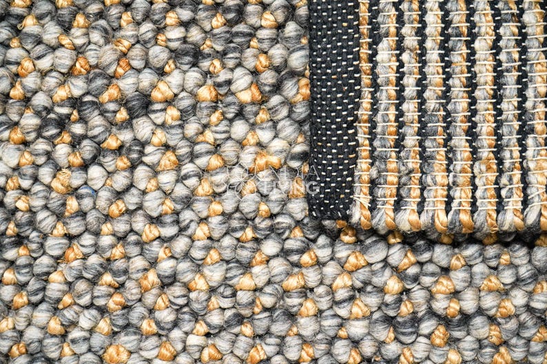 Puede incluir: Una alfombra texturizada con un tejido grueso de lana anudada a mano en una mezcla de tonos gris carb&oacute;n, gris claro y bronceado dorado. La superficie muestra un patr&oacute;n denso y granulado, con una secci&oacute;n que presenta un borde tejido a rayas.