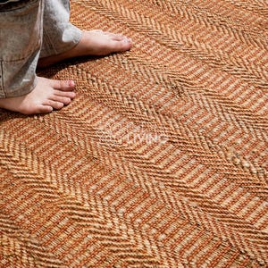 May include: A woven area rug featuring a repeating herringbone pattern in shades of natural tan and burnt orange. The textured surface is constructed from thick, braided fibers, creating a dense and durable floor covering with a distinct zigzag design.