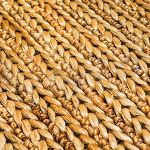 May include: Close-up of a woven rug with a natural, textured appearance. The rug is made of tightly woven, light brown fibers, creating a pattern of diagonal lines. The fibers have variations in color, adding depth and visual interest to the rug.