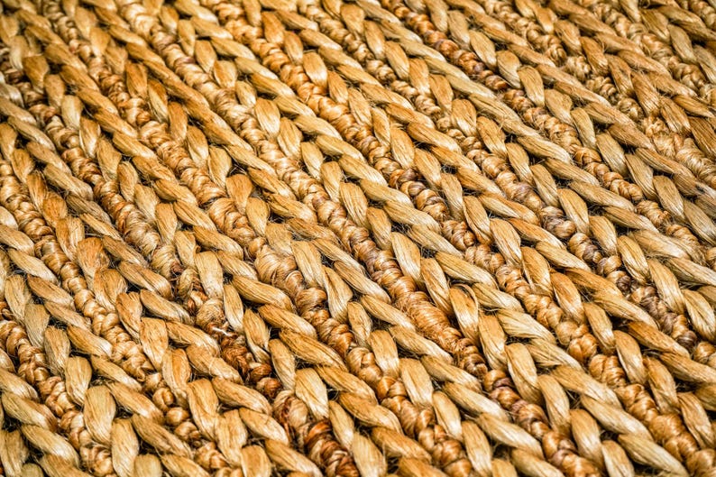 May include: Close-up of a woven rug with a natural, textured appearance. The rug is made of tightly woven, light brown fibers, creating a pattern of diagonal lines. The fibers have variations in color, adding depth and visual interest to the rug.
