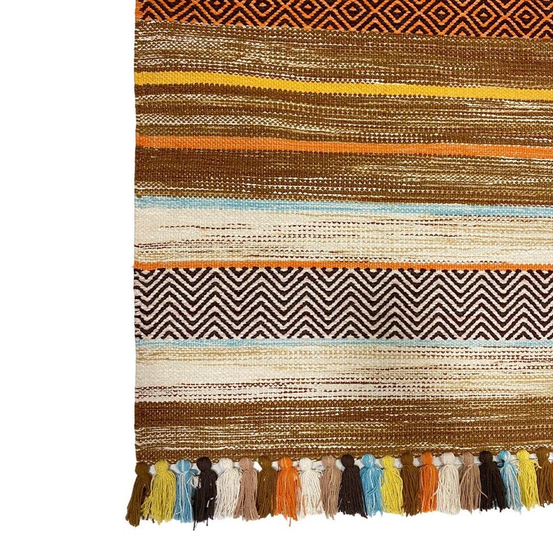 May include: A brown and orange striped rug with a geometric pattern and fringe tassels. The rug has a variety of colors including brown, orange, yellow, blue, and white.