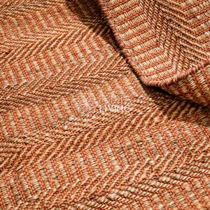 May include: A woven rug featuring a prominent herringbone pattern in alternating shades of burnt orange and natural tan. The texture is thick and fibrous, with a visible weave structure that creates a repeating zigzag design across the surface.