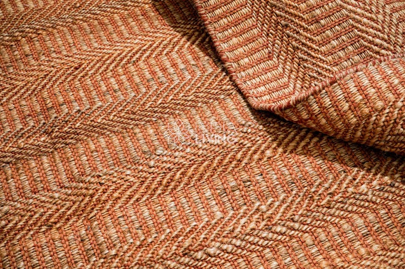 May include: A woven rug featuring a prominent herringbone pattern in alternating shades of burnt orange and natural tan. The texture is thick and fibrous, with a visible weave structure that creates a repeating zigzag design across the surface.