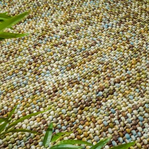 May include: A close-up of a woven rug with a textured surface. The rug is made of small, round, multi-colored beads in shades of brown, green, blue, and white.