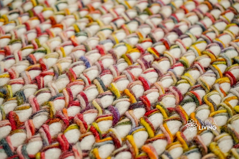 May include: A thick, hand-woven rug featuring a dense, chunky texture made of cream-colored wool yarn interlaced with vibrant, multi-colored strands in red, yellow, green, and purple. The India Living logo is visible in the bottom right corner.