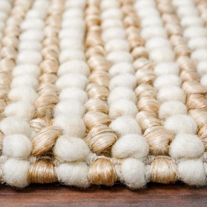 May include: Close-up of a woven rug with a textured surface. The rug has alternating rows of white and beige yarn pom-poms.