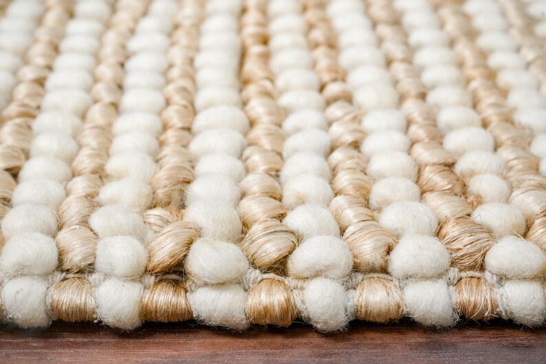 May include: Close-up of a woven rug with a textured surface. The rug has alternating rows of white and beige yarn pom-poms.