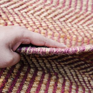 May include: Close-up of a hand holding a woven rug with a chevron pattern in shades of brown and pink. The rug is made of natural fibers and has a textured surface.