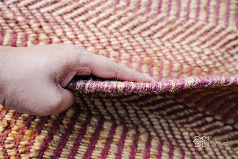 May include: Close-up of a hand holding a woven rug with a chevron pattern in shades of brown and pink. The rug is made of natural fibers and has a textured surface.