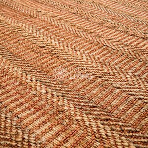 May include: A woven rug featuring a textured herringbone pattern in alternating shades of natural tan and burnt orange. The surface consists of thick, braided fibers arranged in a repeating zigzag design across the entire rectangular area.