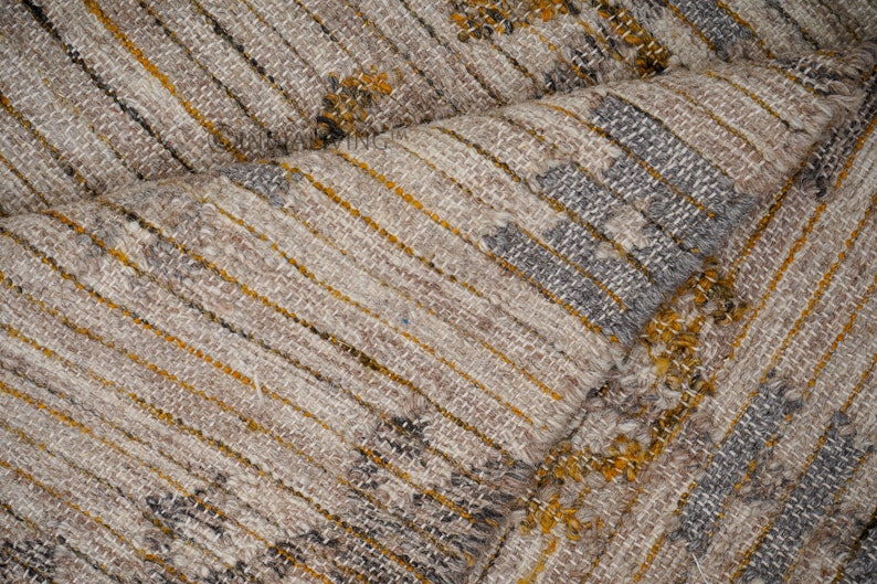 May include: Close-up of a woven fabric with a textured pattern. The fabric is a mix of beige, brown, and gray colors with a subtle yellow stripe.