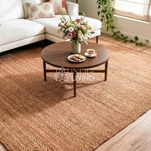 May include: A large rectangular area rug featuring a natural tan color and a woven herringbone pattern. The rug is positioned under a round dark brown wooden coffee table. The text INDIA LIVING is visible in the center of the image.