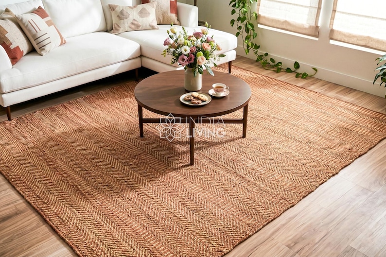 May include: A large rectangular area rug featuring a natural tan color and a woven herringbone pattern. The rug is positioned under a round dark brown wooden coffee table. The text INDIA LIVING is visible in the center of the image.