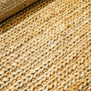 May include: Close-up of a natural fiber rug with a woven texture. The rug is a light tan color with a braided pattern. The rolled-up portion of the rug is visible in the upper left corner.
