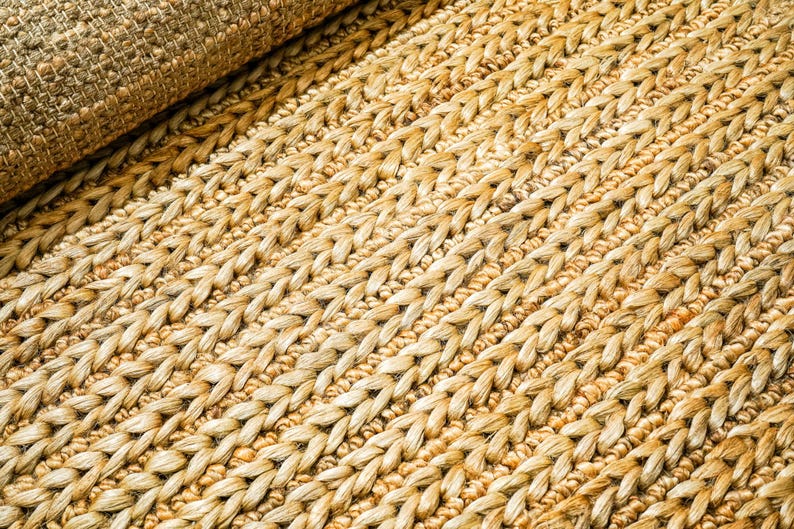 May include: Close-up of a natural fiber rug with a woven texture. The rug is a light tan color with a braided pattern. The rolled-up portion of the rug is visible in the upper left corner.