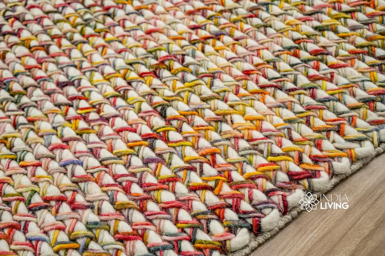May include: A thick, hand-woven area rug featuring a chunky braided texture. The design consists of cream-colored yarn interwoven with vibrant multi-colored strands in shades of red, yellow, blue, and green. The India Living logo is visible.