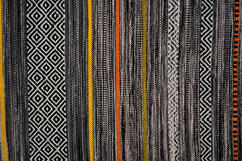 Op de afbeelding: Een close-up van een geweven tapijt met verschillende zwarte, witte, grijze, gele en oranje strepen. Het tapijt heeft een geometrisch patroon.