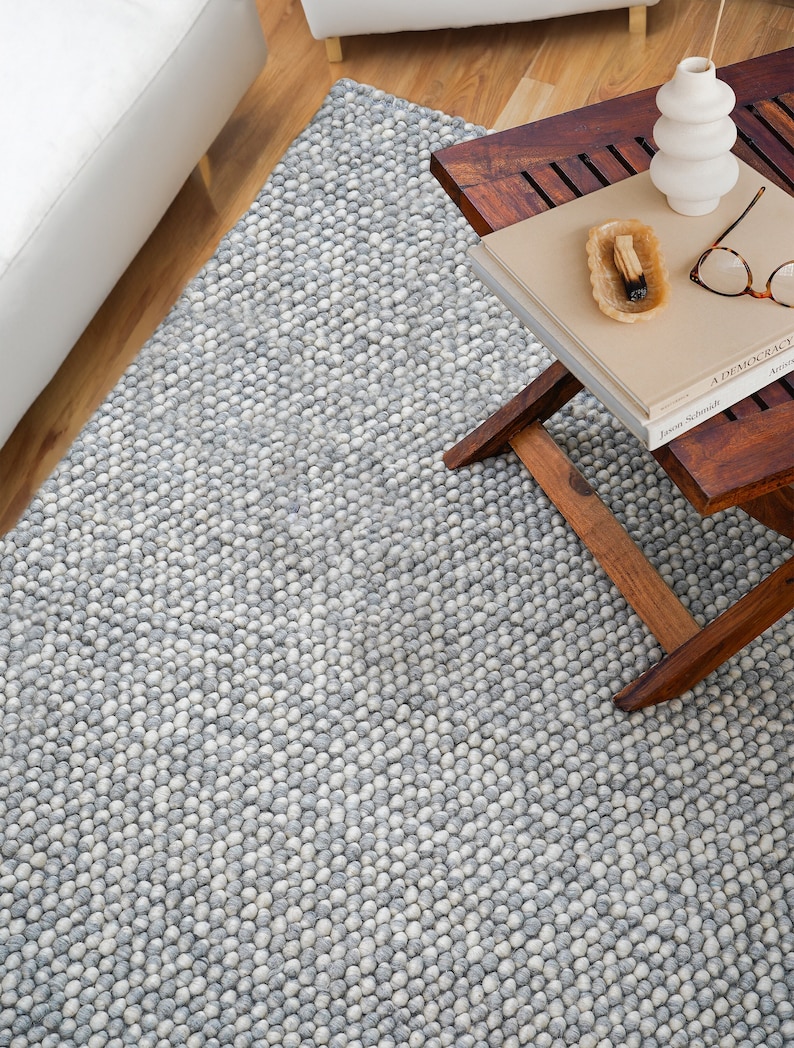 May include: A gray and white round wool rug with a textured surface. The rug is made of small, round balls of wool that are tightly packed together. The rug is shown in a room with a wooden floor and a wooden table with a book and a vase on it.