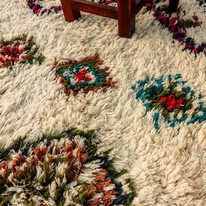 Op de afbeelding: Een wit, pluche tapijt met een geometrisch patroon in tinten rood, blauw, groen en bruin. Het tapijt heeft een verweerde, vintage uitstraling.