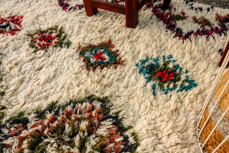 Op de afbeelding: Een wit, pluche tapijt met een geometrisch patroon in tinten rood, blauw, groen en bruin. Het tapijt heeft een verweerde, vintage uitstraling.