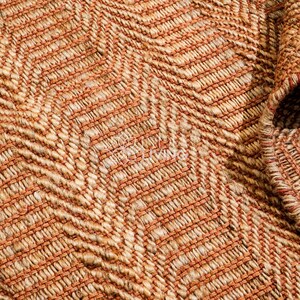 May include: A woven rug featuring a textured chevron pattern in shades of tan, beige, and burnt orange. The material consists of thick, interlaced fibers creating a dense, raised surface with a visible border edge folded over at the corner.