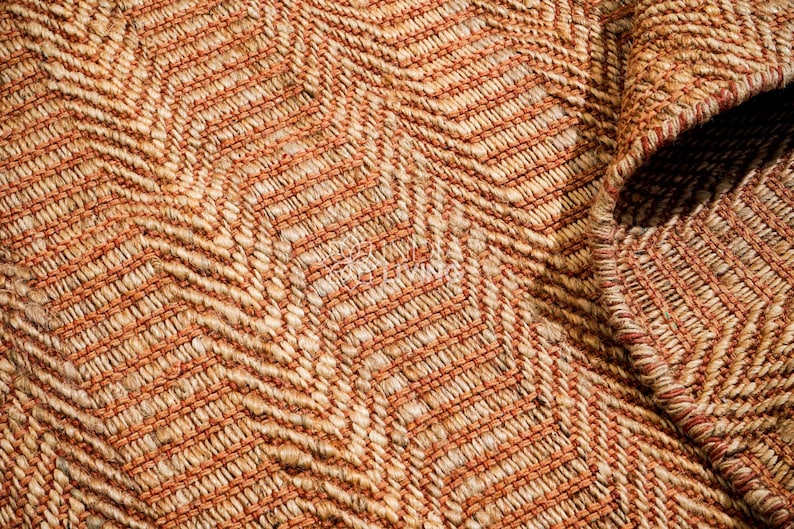 May include: A woven rug featuring a textured chevron pattern in shades of tan, beige, and burnt orange. The material consists of thick, interlaced fibers creating a dense, raised surface with a visible border edge folded over at the corner.