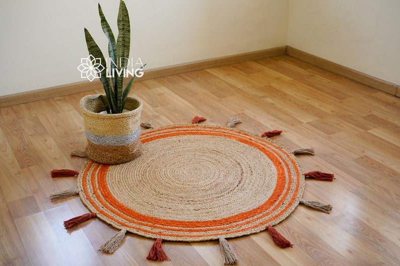 May include: A round jute rug with orange stripes and tassels around the edge. The rug is on a wooden floor and a potted plant is in the background.