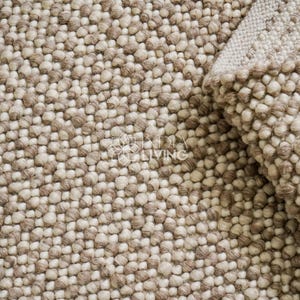 Könnte beinhalten: Nahaufnahme eines strukturierten Teppichs mit einem sich wiederholenden Muster aus kleinen, abgerundeten, beige- und braunen Elementen. Der Teppich ist teilweise gerollt und zeigt eine hellere beige Unterseite. Das Design deutet auf einen gemütlichen Einrichtungsgegenstand in neutralen Tönen hin.
