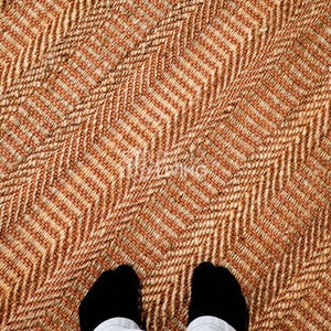 May include: A woven rug featuring a prominent herringbone pattern in alternating shades of tan, beige, and light brown. The texture appears coarse and fibrous, with a consistent diagonal orientation across the entire surface of the floor covering.