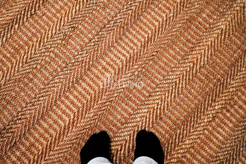 May include: A woven rug featuring a prominent herringbone pattern in alternating shades of tan, beige, and light brown. The texture appears coarse and fibrous, with a consistent diagonal orientation across the entire surface of the floor covering.