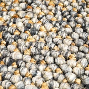 Puede incluir: Una alfombra gruesa tejida a mano con una textura densa de fibras redondeadas y anudadas. La superficie se compone de una paleta de colores variados que incluye tonos gris carb&oacute;n, gris claro, crema y bronceado c&aacute;lido, creando un aspecto tonal.