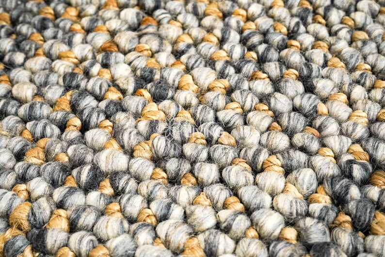 Puede incluir: Una alfombra gruesa tejida a mano con una textura densa de fibras redondeadas y anudadas. La superficie se compone de una paleta de colores variados que incluye tonos gris carb&oacute;n, gris claro, crema y bronceado c&aacute;lido, creando un aspecto tonal.