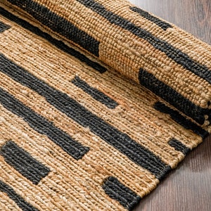 Puede incluir: Una alfombra de yute enrollada con rayas negras y de color natural. La alfombra tiene un patrón geométrico.