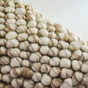 K&ouml;nnte beinhalten: Nahaufnahme eines beige-braunen Wollteppichs mit einer strukturierten, blasenartigen Oberfl&auml;che. Der Teppich besteht aus kleinen, runden Wollkugeln, die miteinander verwoben sind.