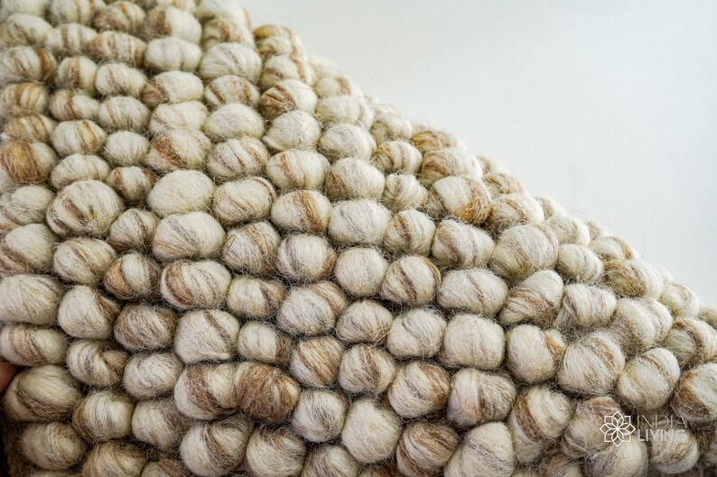 K&ouml;nnte beinhalten: Nahaufnahme eines beige-braunen Wollteppichs mit einer strukturierten, blasenartigen Oberfl&auml;che. Der Teppich besteht aus kleinen, runden Wollkugeln, die miteinander verwoben sind.