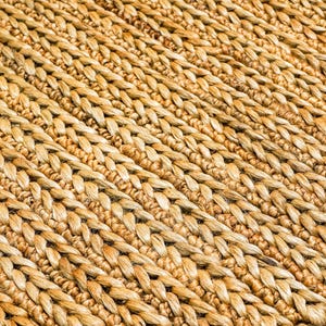 May include: Close-up of a woven rug with a natural, light brown color. The rug has a textured surface with a diagonal pattern created by the woven fibers. The fibers are tightly woven together, creating a durable and visually appealing texture.