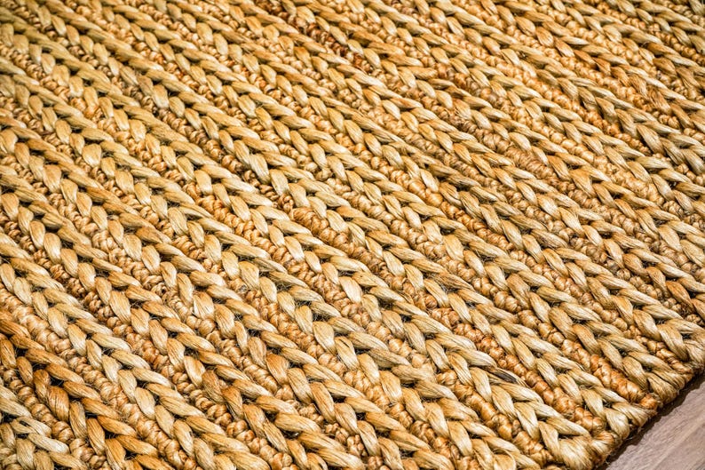 May include: Close-up of a woven rug with a natural, light brown color. The rug has a textured surface with a diagonal pattern created by the woven fibers. The fibers are tightly woven together, creating a durable and visually appealing texture.