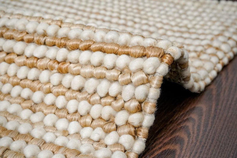 May include: Close-up of a beige and white woven rug with a textured, bumpy surface. The rug is made of natural fibers and has a contemporary design.