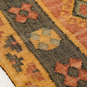 Puede incluir: Primer plano de una alfombra tejida con un patr&oacute;n geom&eacute;trico en tonos marrones, naranjas y verdes. La alfombra tiene una superficie texturizada y un borde de flecos.