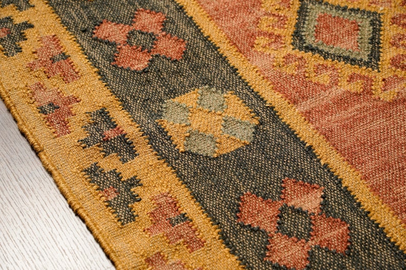 Puede incluir: Primer plano de una alfombra tejida con un patr&oacute;n geom&eacute;trico en tonos marrones, naranjas y verdes. La alfombra tiene una superficie texturizada y un borde de flecos.