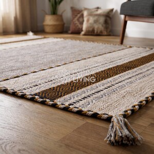 May include: A rectangular area rug featuring a woven pattern of cream, tan, brown, and black stripes with geometric motifs. The rug has a thick braided border and a corner tassel made of twisted fibers in matching colors, with a logo in the center.