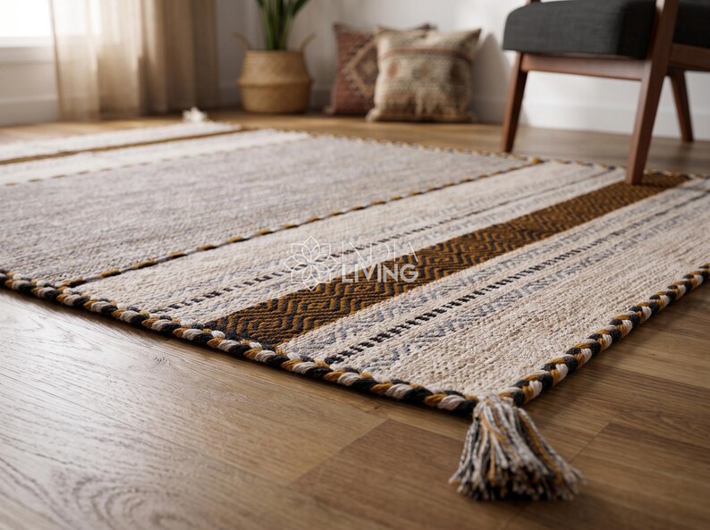 May include: A rectangular area rug featuring a woven pattern of cream, tan, brown, and black stripes with geometric motifs. The rug has a thick braided border and a corner tassel made of twisted fibers in matching colors, with a logo in the center.