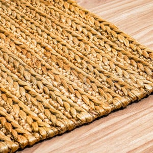 May include: Close-up of a woven rug with a natural, light brown color. The rug has a textured, braided design and is placed on a wooden surface with visible wood grain.