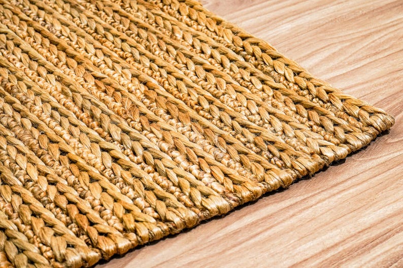 May include: Close-up of a woven rug with a natural, light brown color. The rug has a textured, braided design and is placed on a wooden surface with visible wood grain.