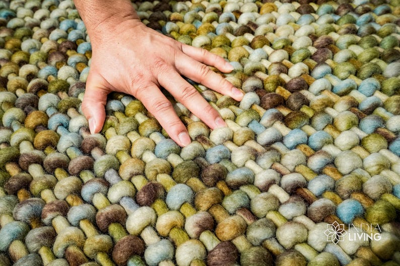 May include: Close-up of a hand resting on a woven rug with a textured, pebble-like design. The rug is made of wool in shades of green, brown, and blue.